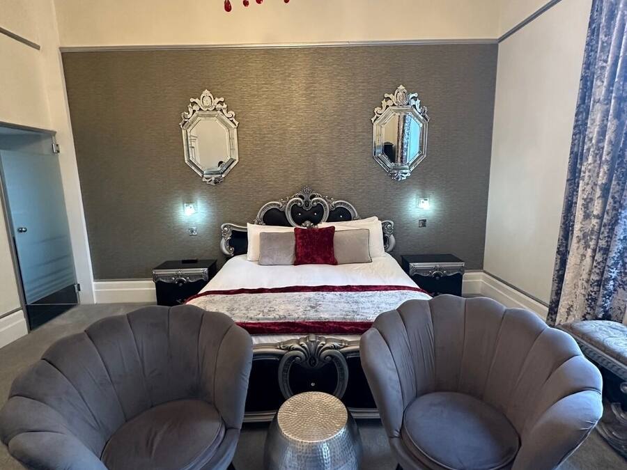 A bedroom with a bed, two armchairs, a chandelier, and two wall mirrors.