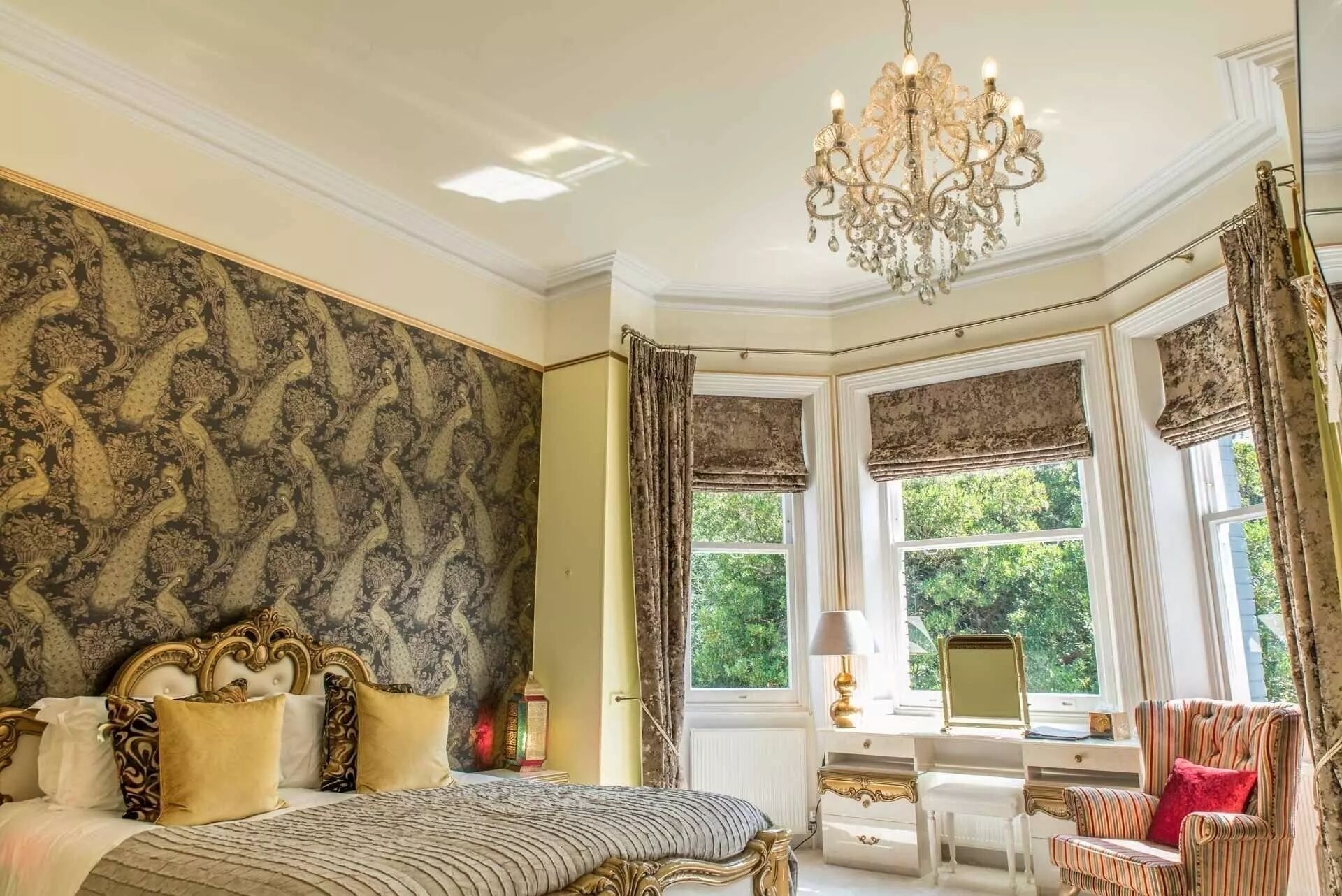A luxurious bedroom with a large bed, ornate wallpaper, a chandelier, and a view of greenery through the window.