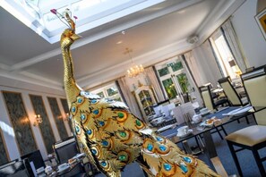 A dining area with tables and chairs, a chandelier, and a decorative peacock statue. - Derby Manor (Bournemouth)