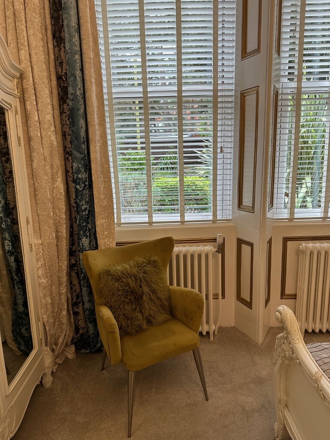 A room with a yellow armchair, a large window with blinds, a radiator, and a white ornate bed.
