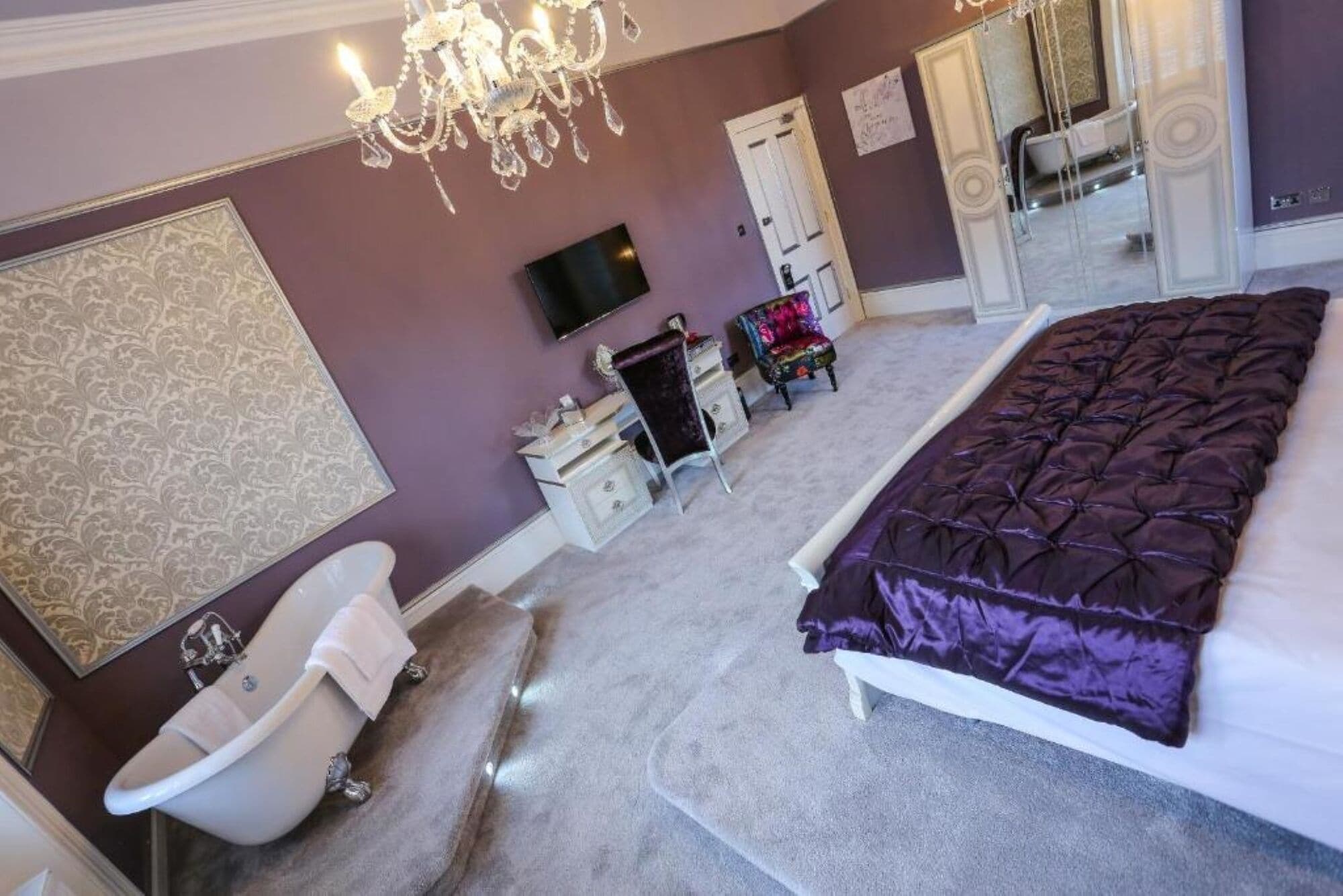 A bedroom with a large bed, a white bathtub, a television, and a chandelier.