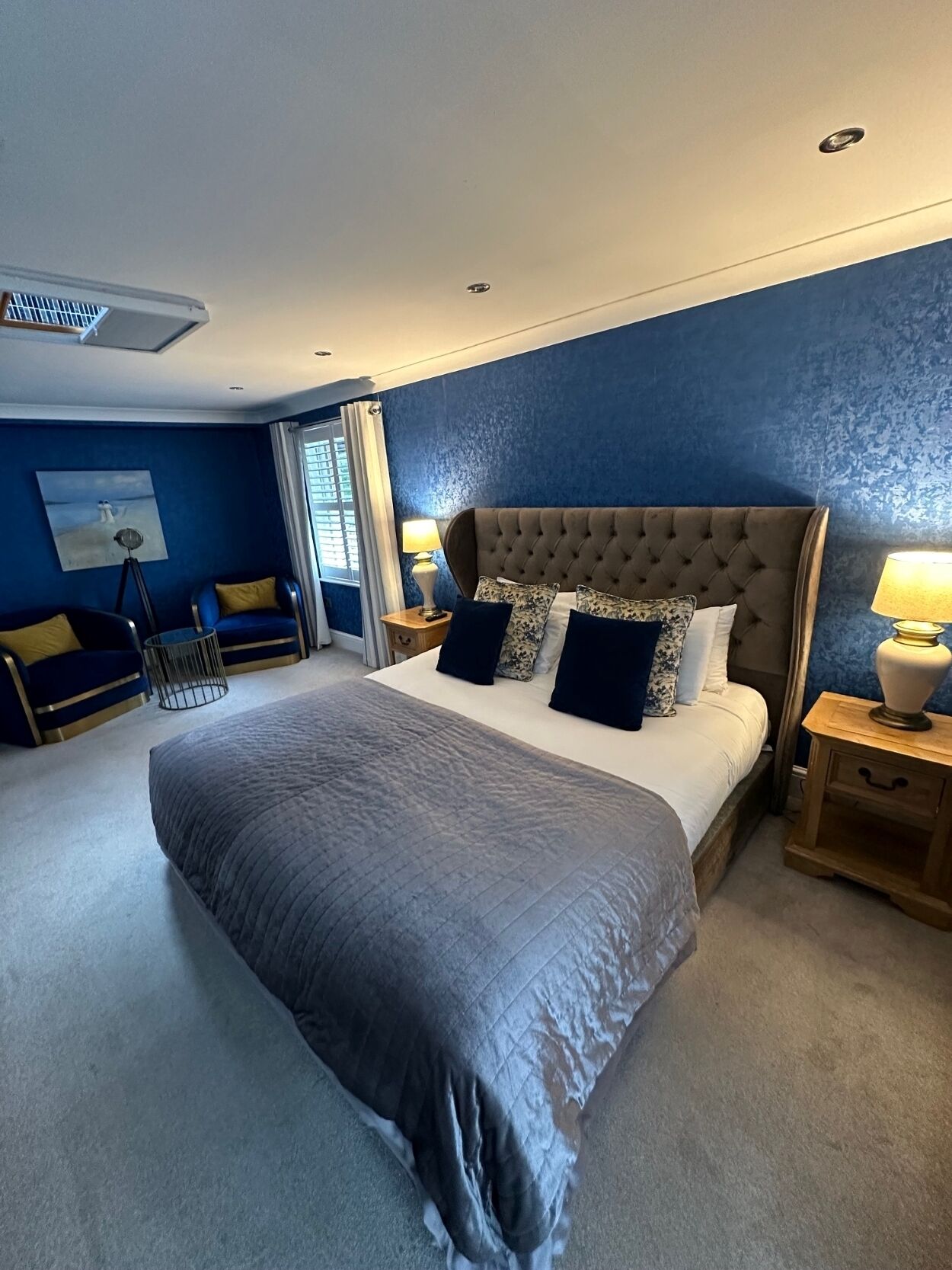 A spacious bedroom with a large bed, a nightstand, a lamp, and a window with blinds.