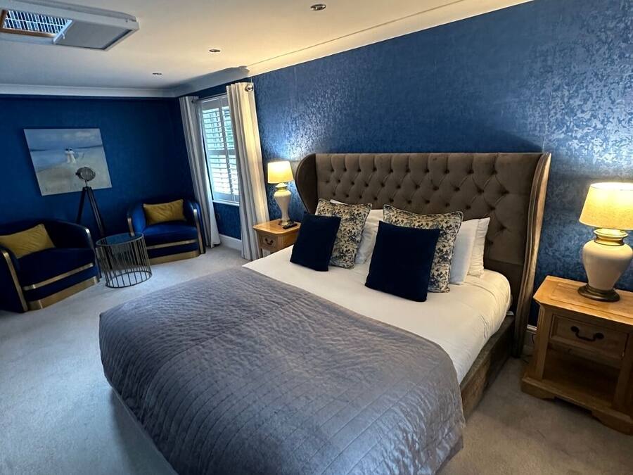 A spacious bedroom with a large bed, a nightstand, a lamp, and a window with blinds.