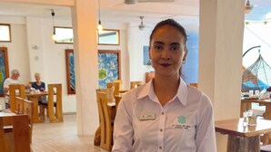 A person in a white shirt and apron holding a tray with food, standing in a restaurant with wooden furniture and a tiled floor. - Hotel La Casa de Judy (Puerto Ayora)