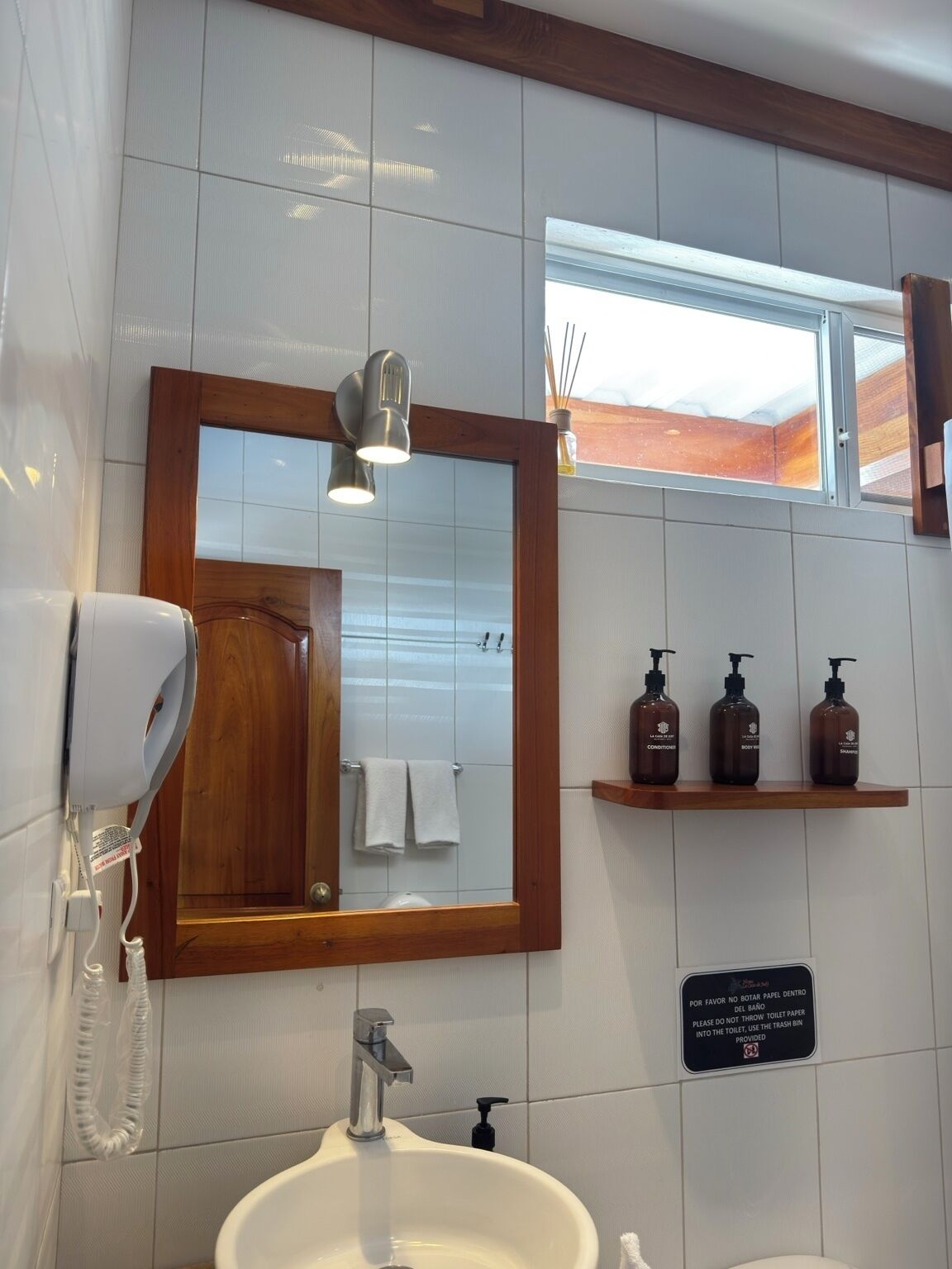 A bathroom with a mirror, a hairdryer, a sink, and toiletries on a shelf.