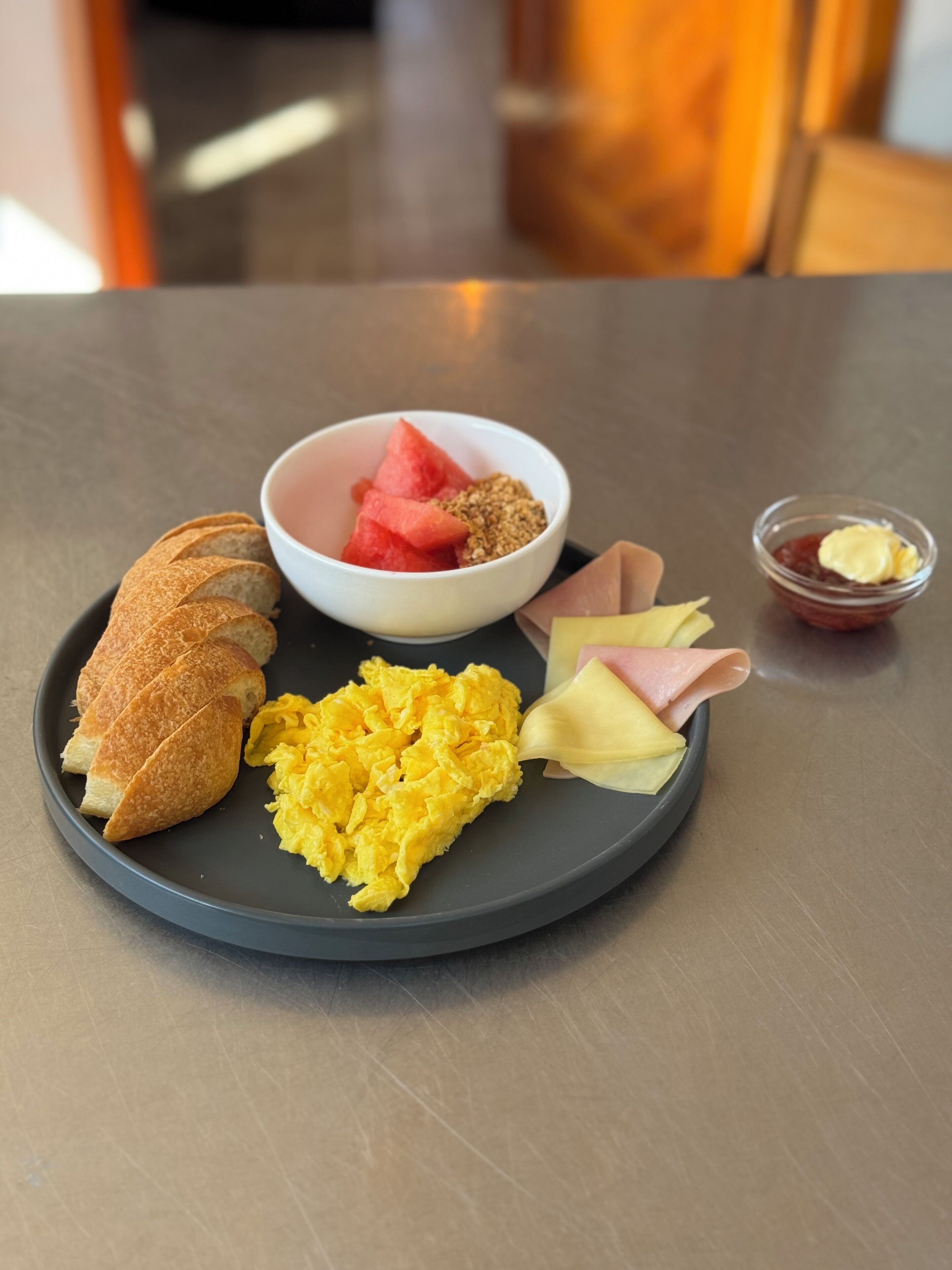 A plate with scrambled eggs, watermelon, granola, ham, and a croissant on a table.