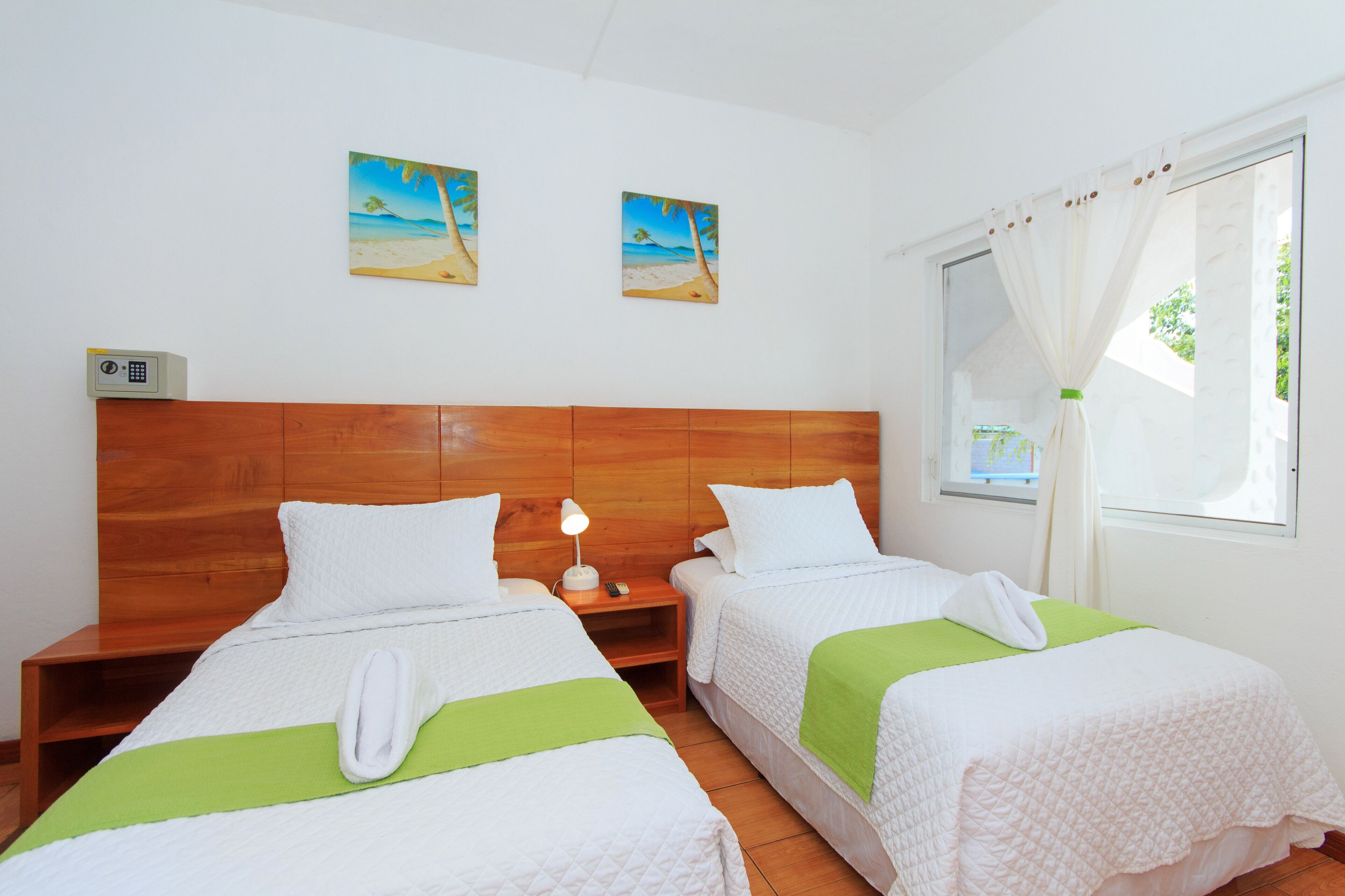 A hotel room with two beds, a wooden headboard, a nightstand with a lamp, and two beach-themed wall art pieces.