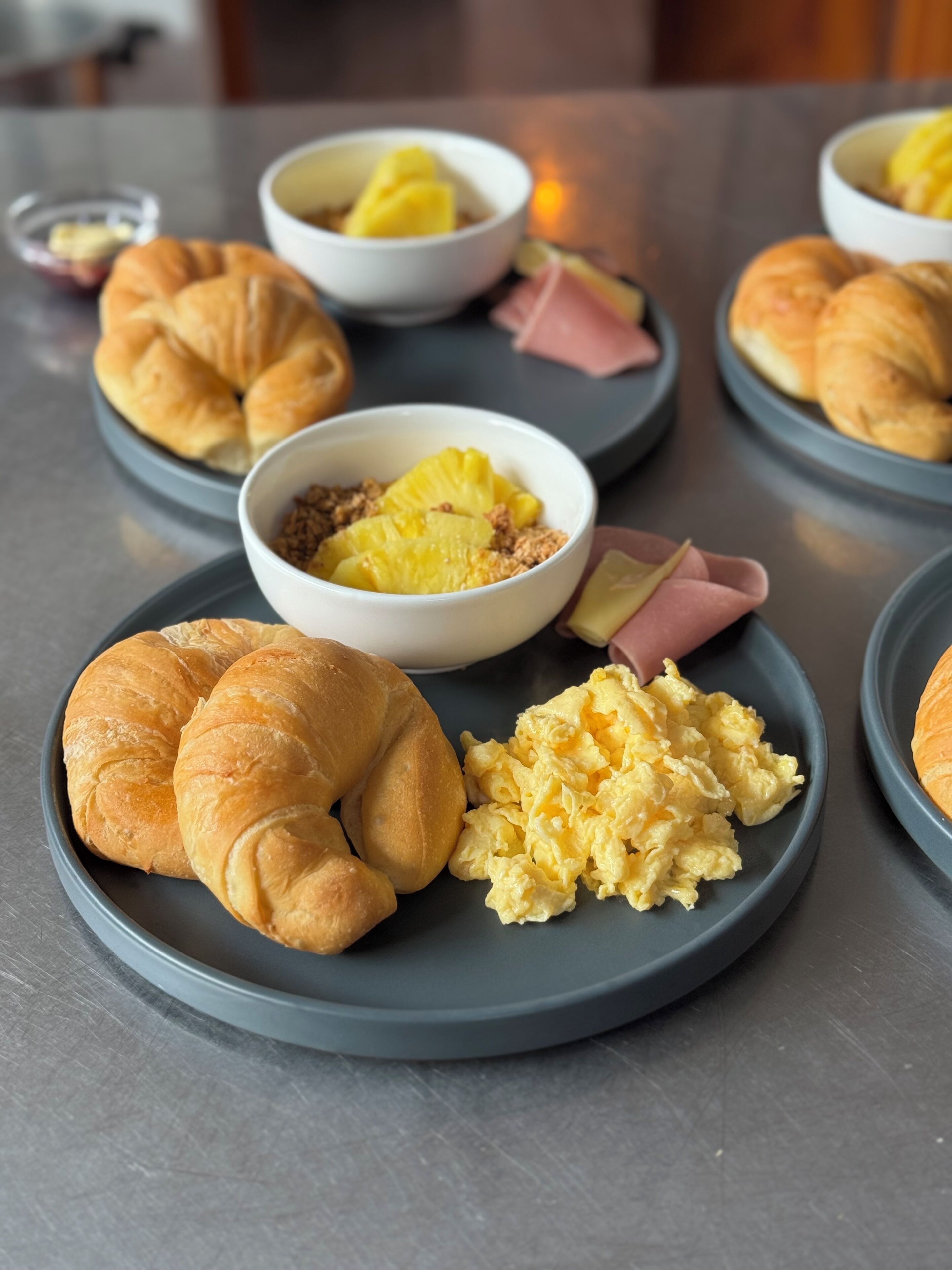 Desayuno con croissants, huevos revueltos y un cuenco de fruta en platos.