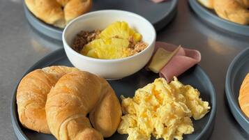 Desayuno con croissants, huevos revueltos y un cuenco de fruta en platos.