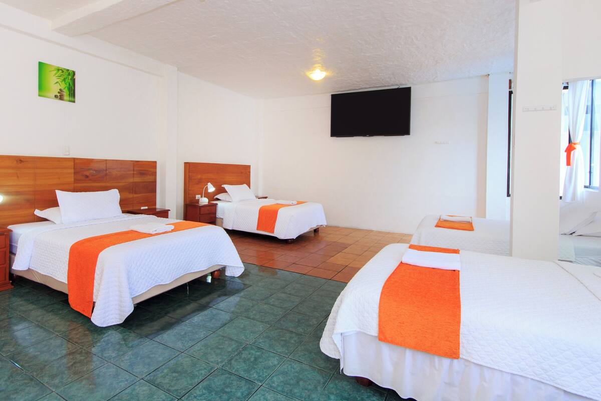 A hotel room with three beds, each with white bedding and orange accents. There is a wooden headboard on the left bed, a flat-screen TV mounted on the wall, and a green tiled floor.