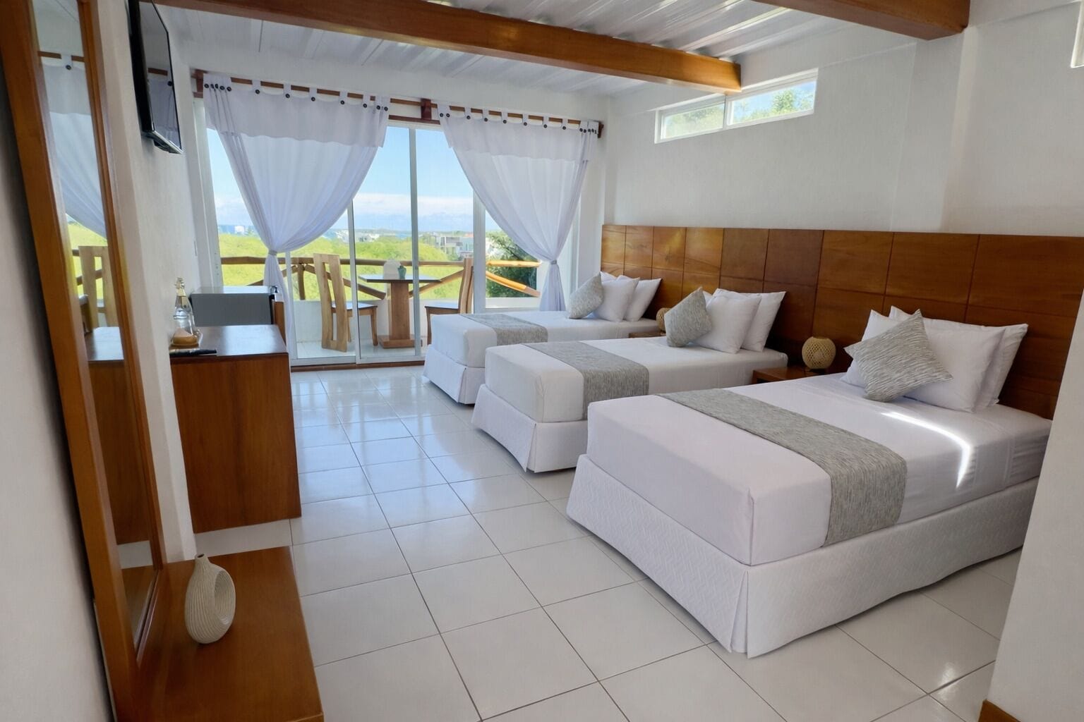 Habitación de hotel con dos camas, cabecero de madera, zona de comedor con mesa y sillas, y vistas a través de amplios ventanales.