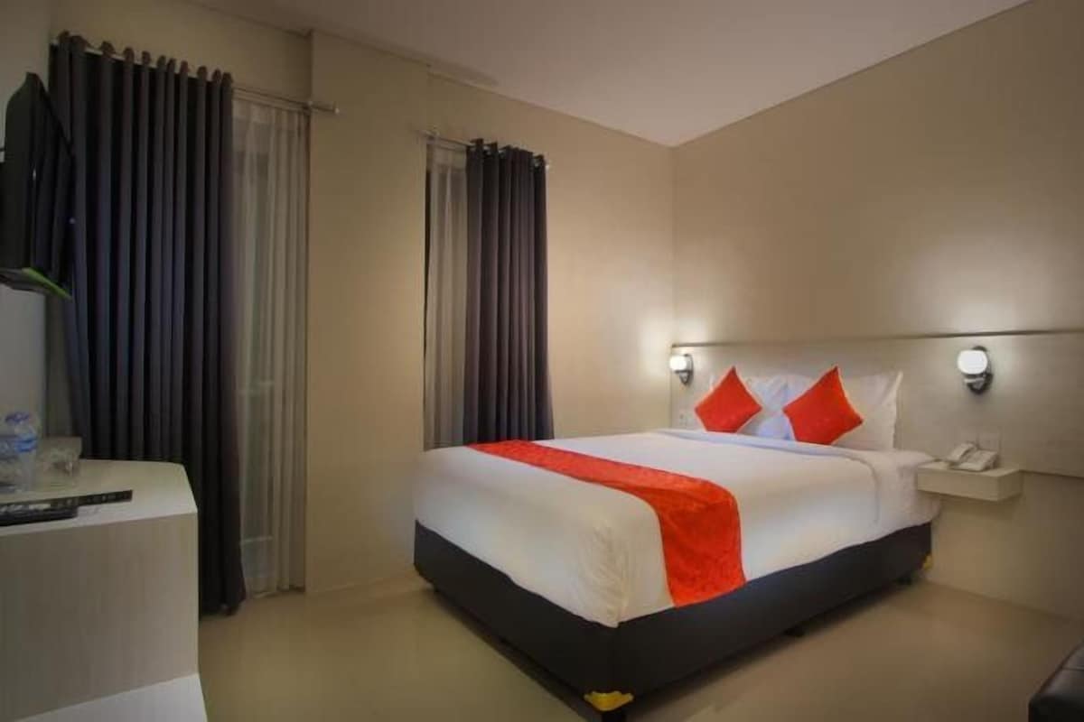 A hotel room with a bed, a bedside table, a nightstand, a lamp, and a window with curtains.