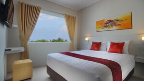 A hotel room with a large bed, beige curtains, a window with a view, a painting on the wall, and a small table. - The Rinaya Canggu (Tibubeneng)