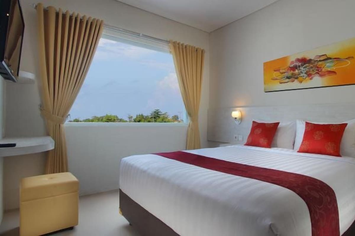 A hotel room with a large bed, beige curtains, a window with a view, a painting on the wall, and a small table.