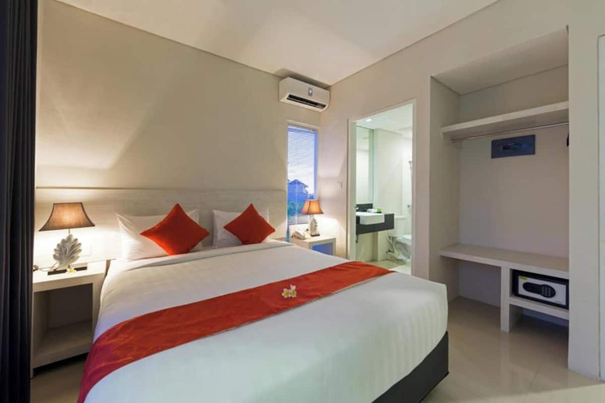 A hotel room with a bed, nightstands, a desk, an air conditioning unit, a wardrobe, and a bathroom visible through an open door.