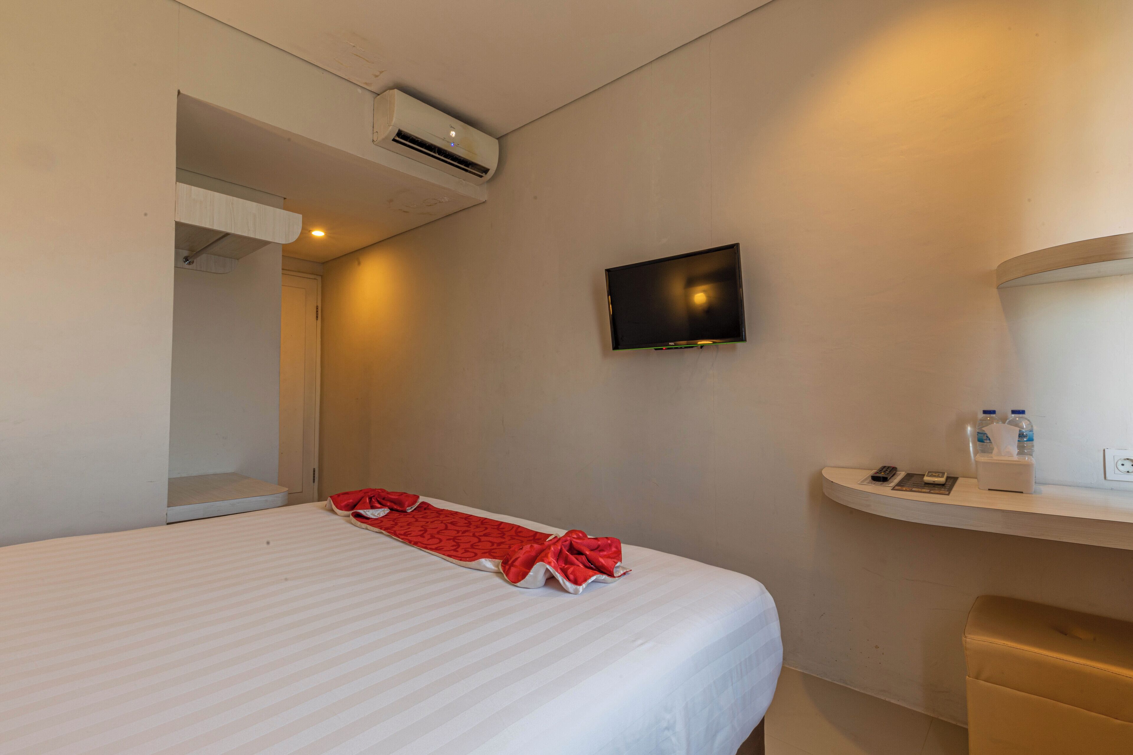 A hotel room with a bed, a mounted TV, a bedside table with a phone and a tissue box, and an air conditioning unit.