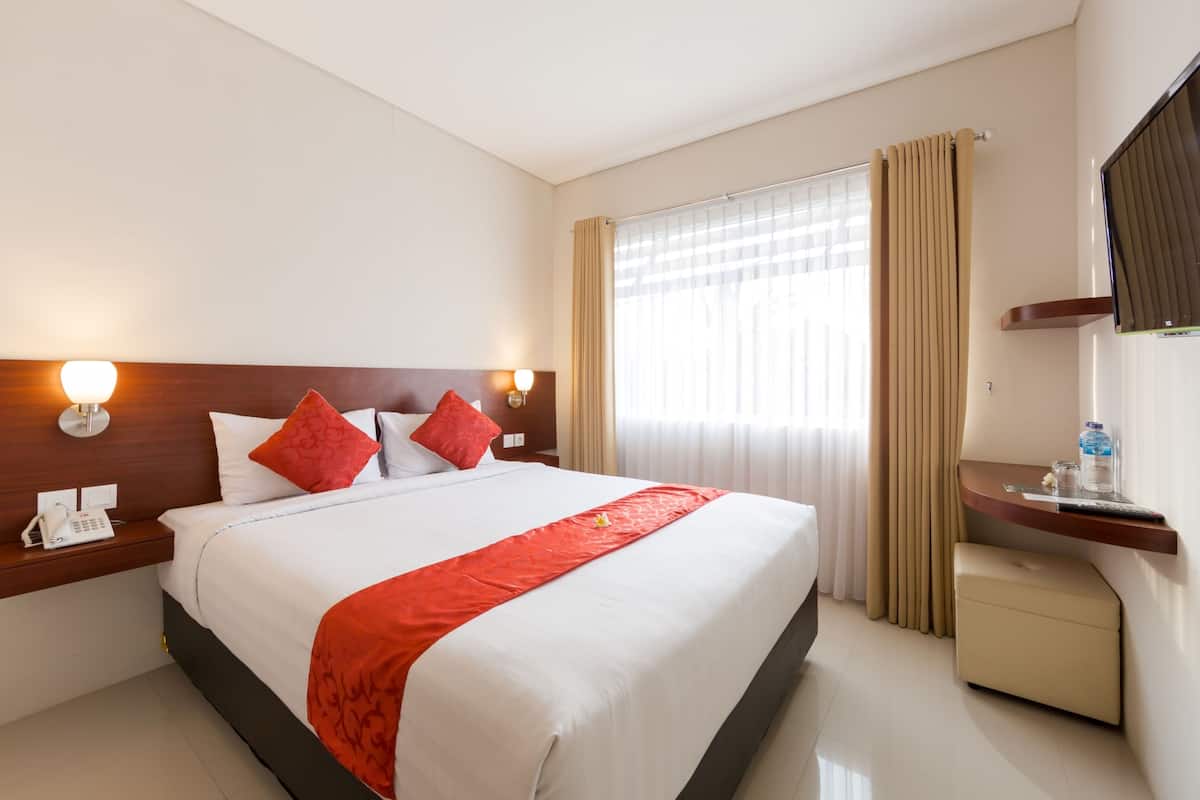 A hotel room with a large bed, two bedside tables with lamps, a small bedside table with a phone, a nightstand, a window with curtains, and a small table with a bottle and glasses.