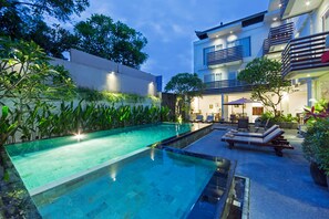 Une piscine dotée d’un espace salon en contrebas, entourée d’une végétation luxuriante et bordée d’un bâtiment moderne avec des balcons.