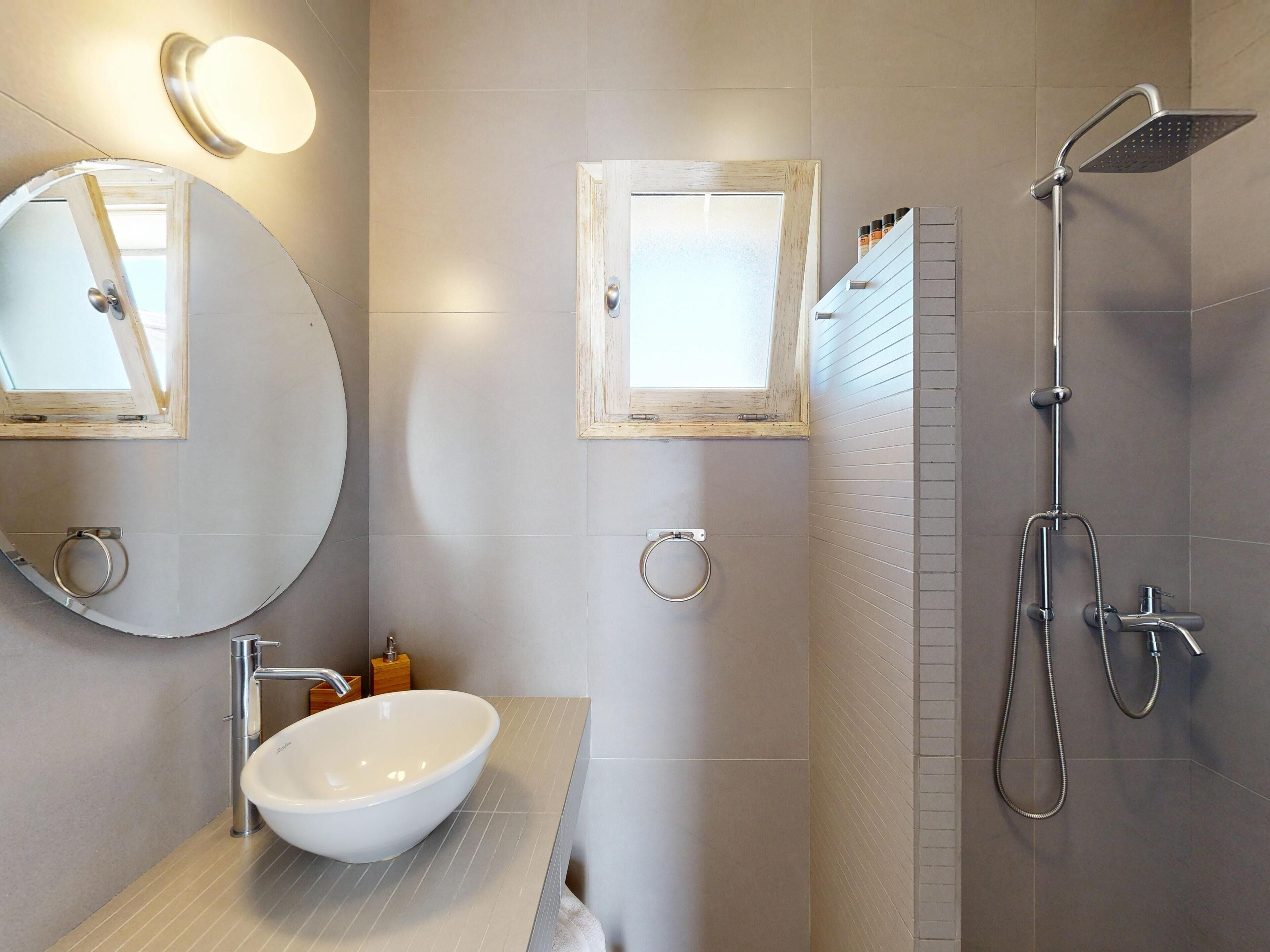 Un baño moderno con un espejo grande y redondo, un espejo rectangular, un lavabo blanco, un cabezal de ducha de mano y un perchero.