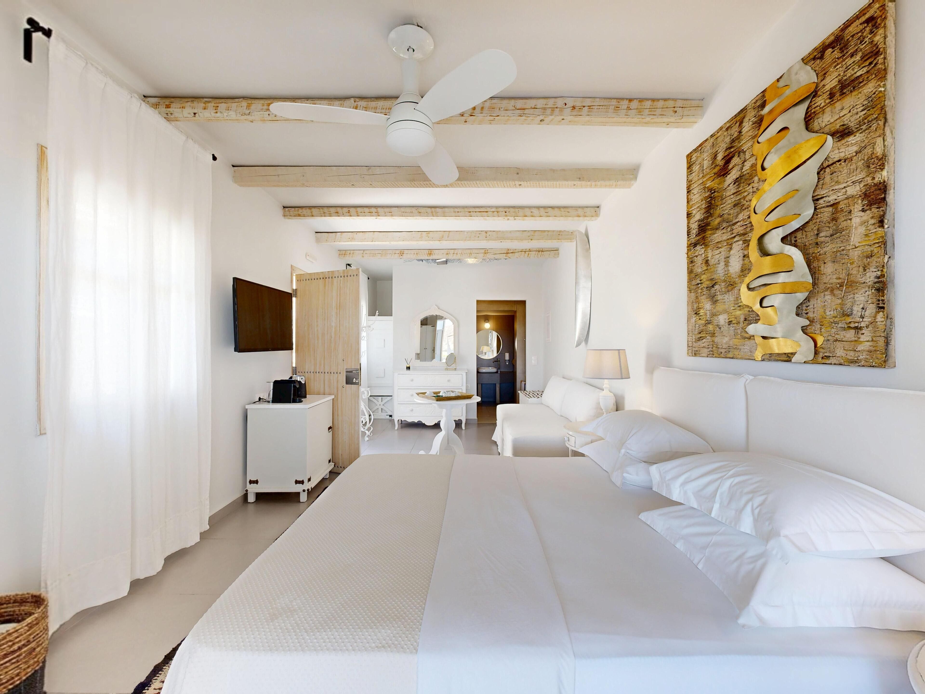 Une chambre d’hôtel spacieuse, dotée d’un grand lit, d’un ventilateur de plafond, d’un plafond en bois, d’une commode blanche, d’une coiffeuse avec un miroir et d’une œuvre d’art accrochée au mur.