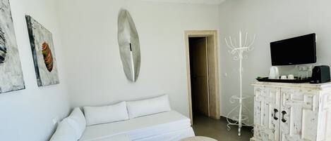 Un dormitorio moderno y minimalista con un sofá blanco, un ventilador de techo, un televisor montado y un mueble blanco con una bandeja que contiene una cafetera y tazas.