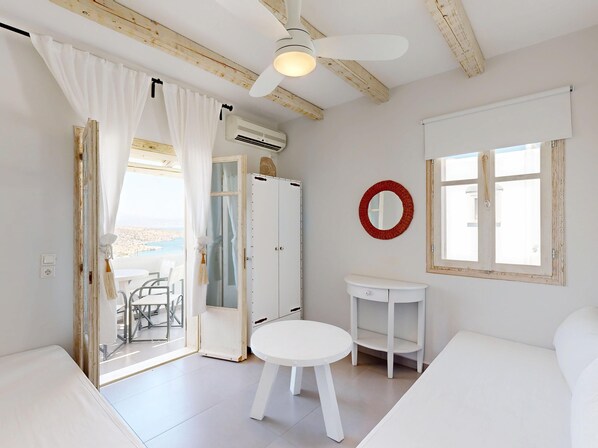 A room with a bed, a round mirror, a white table, a white bench, a ceiling fan, and a view of the beach through an open door. - Speires (Naxos)
