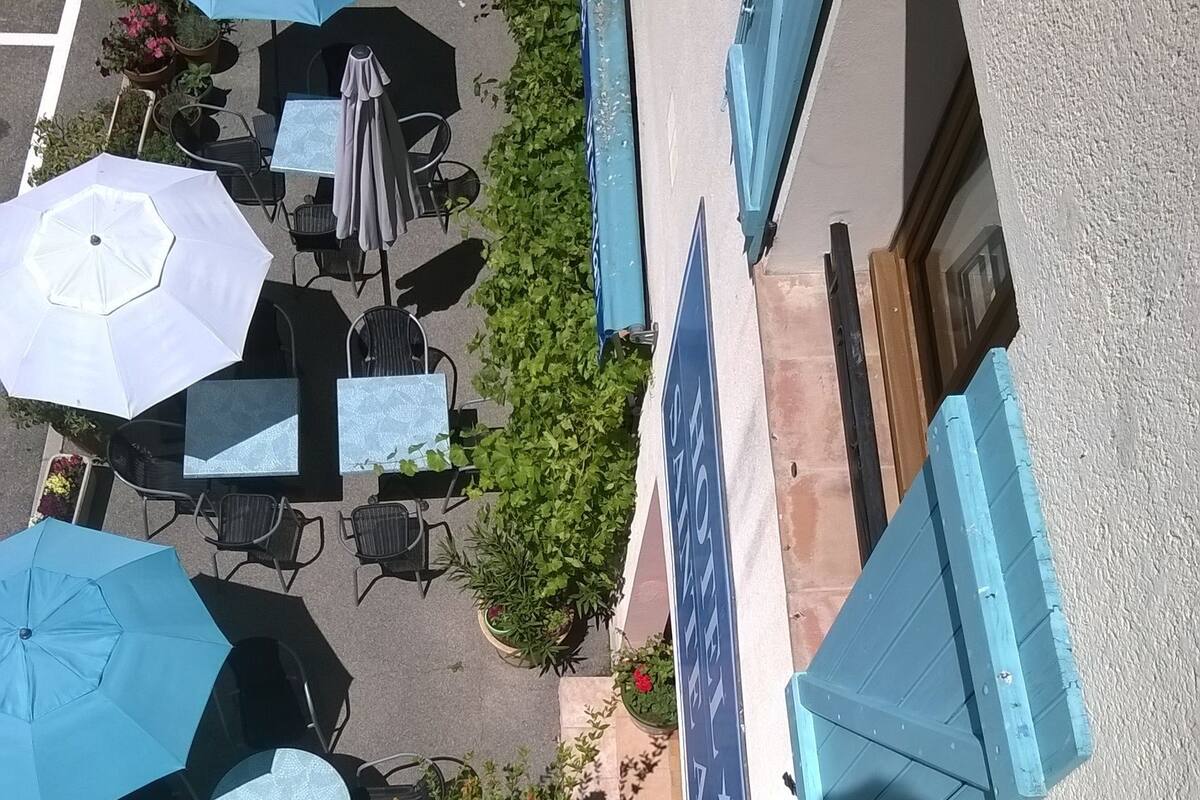 Ein Außenbereich mit Tischen und blauen Sonnenschirmen, umgeben von Grünpflanzen und einem Gebäude mit blauem Vordach.