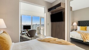 Habitación de hotel con una cama grande, un televisor montado en la pared, un escritorio con una silla y vistas a la ciudad a través de amplios ventanales.