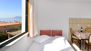 Una habitación pequeña y luminosa con una cama, una mesa con una botella de vino y vistas al mar y a las montañas.