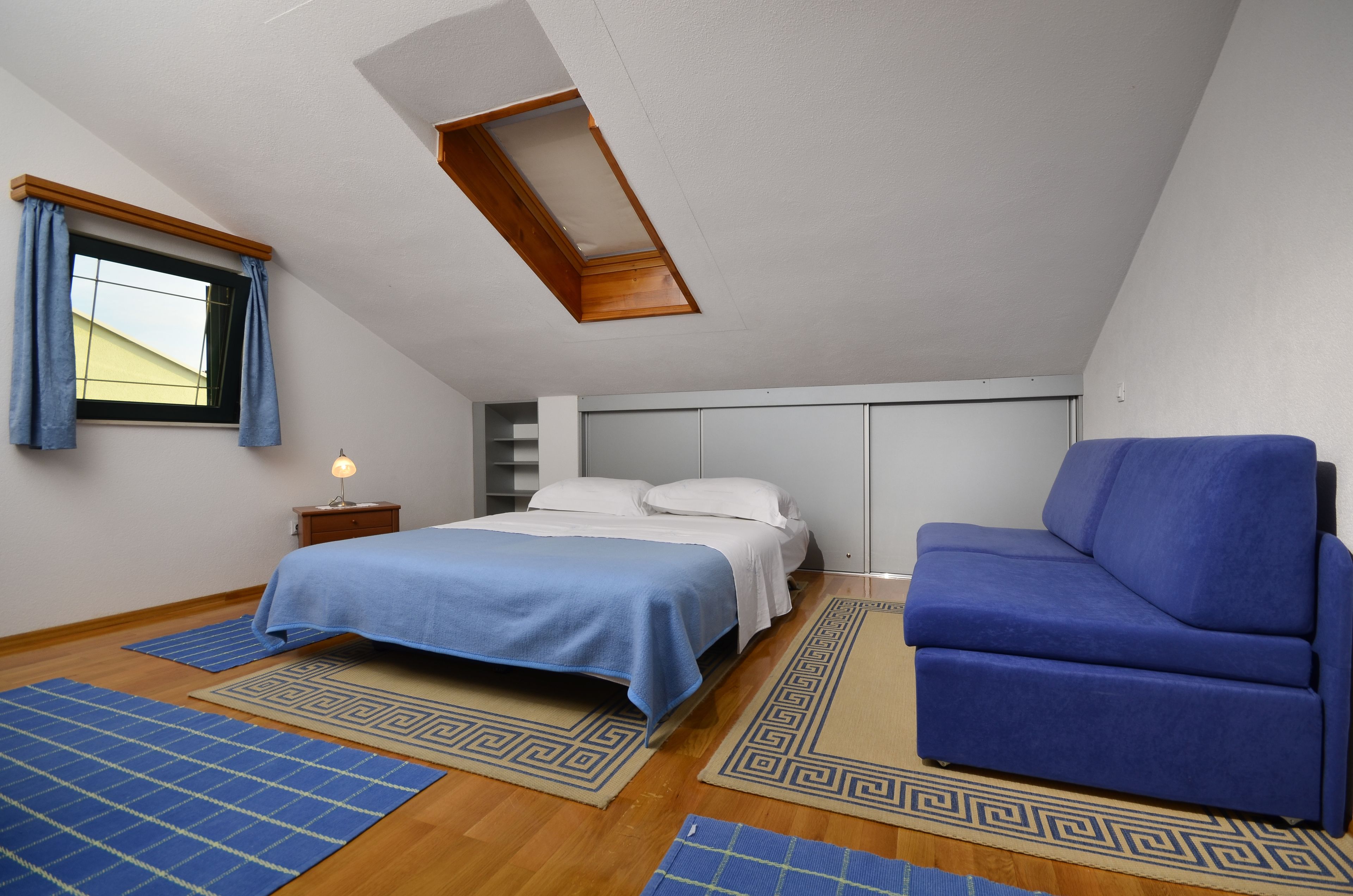 A bedroom with a bed, a blue chair, a nightstand with a lamp, and a skylight.
