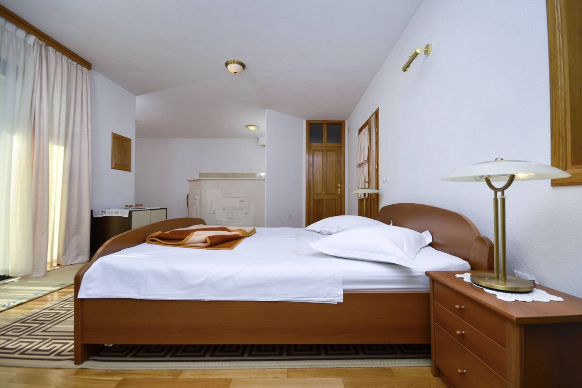 Un lit bien fait, recouvert d’une couette blanche et d’un plaid marron, une table de chevet en bois avec une lampe, et une commode en bois avec un miroir.