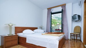 Una cama bien hecha con sábanas blancas, una mesita de noche de madera con una lámpara, una silla y un televisor montado en la pared.