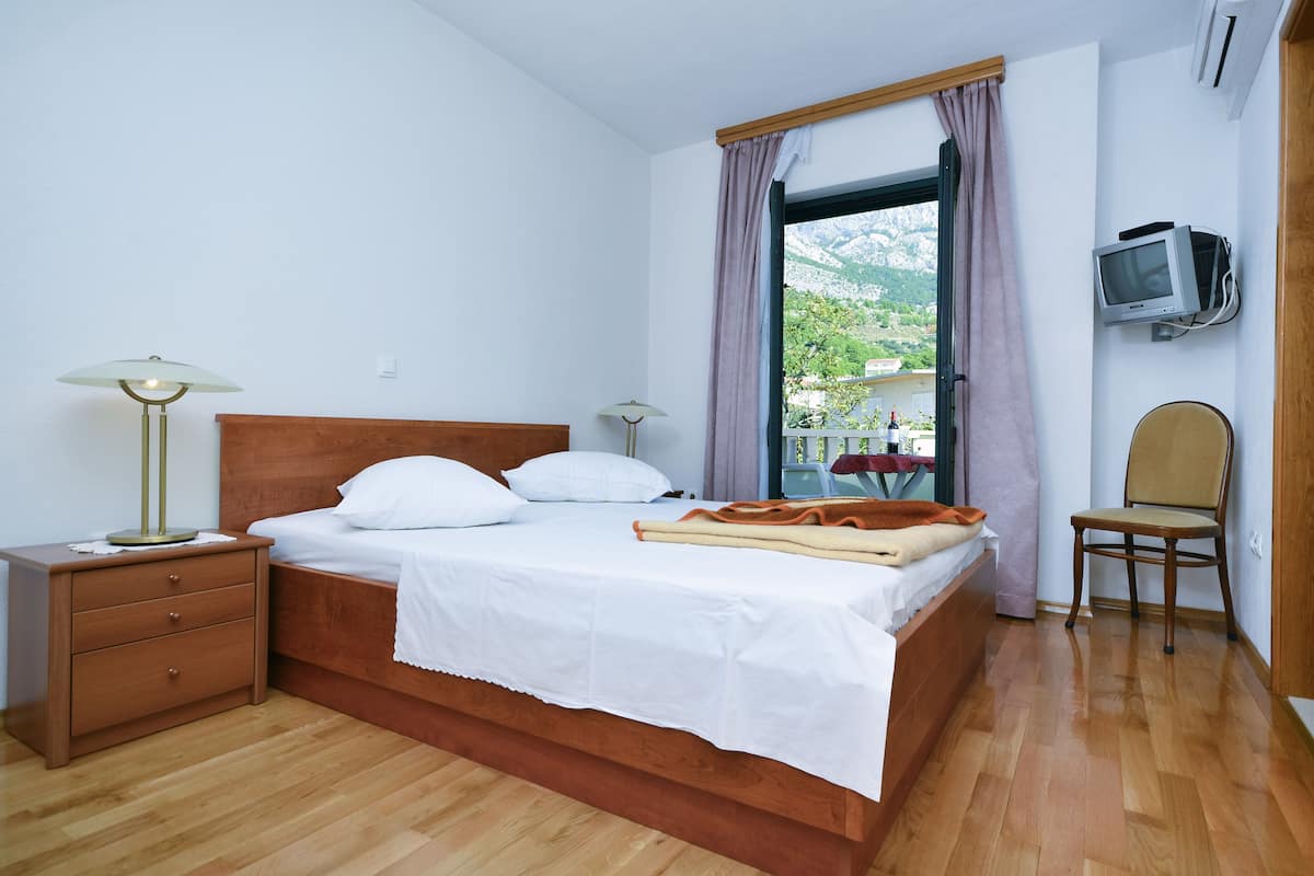 Un lit bien fait, avec du linge de lit blanc, une table de chevet en bois sur laquelle se trouve une lampe, une chaise et un téléviseur fixé au mur.