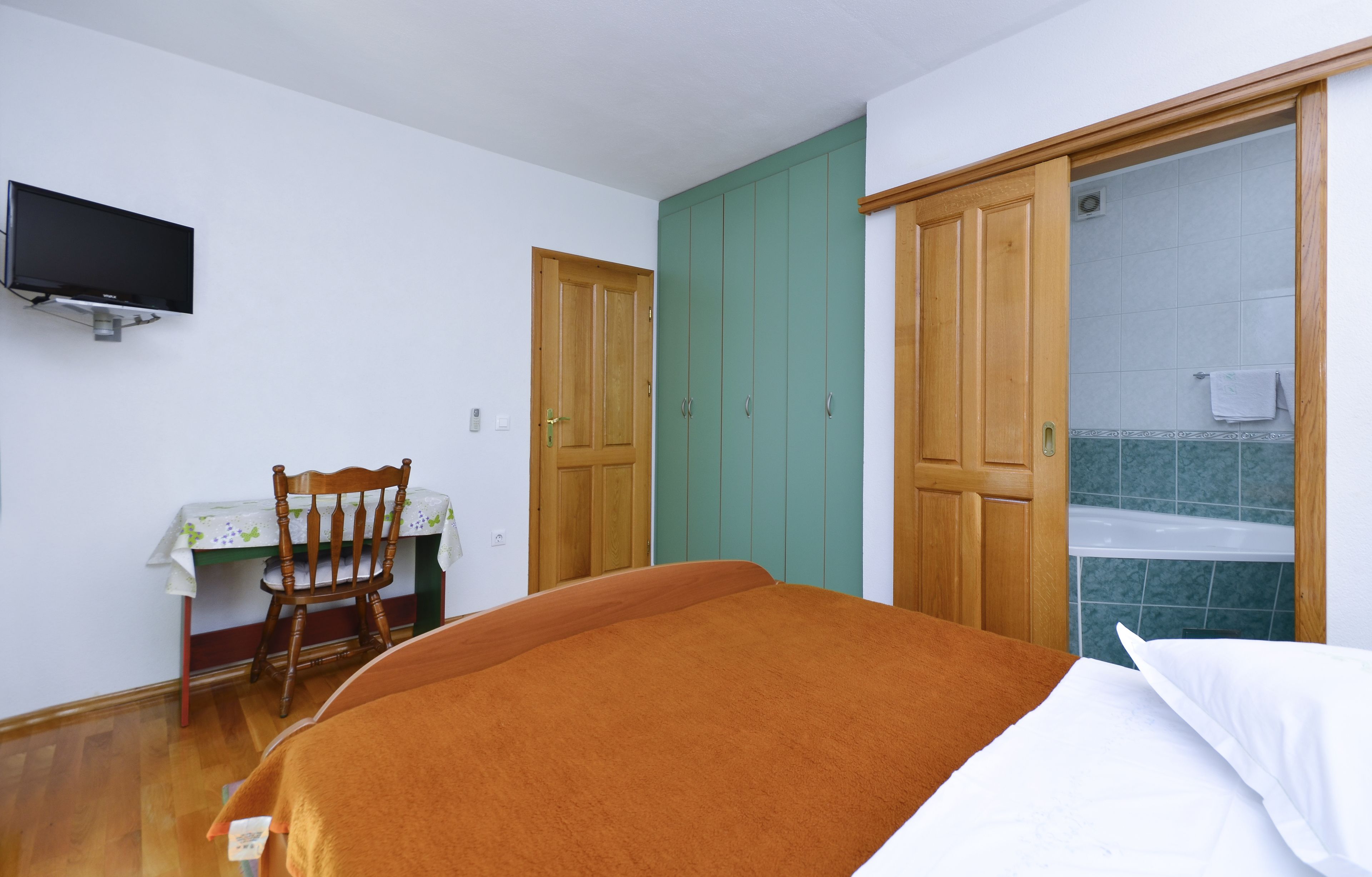 Un dormitorio con cama, un armario de madera, un televisor montado en la pared y una mesita de noche con una silla.