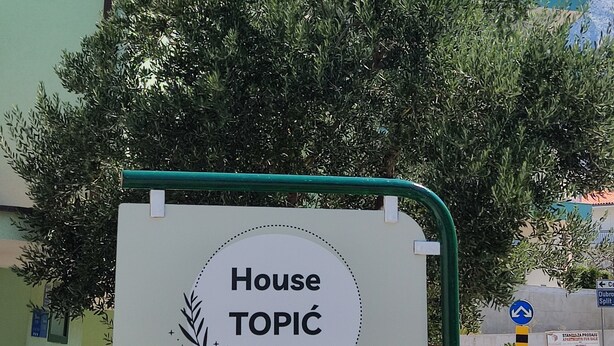 Letrero que dice "House TOPIC", dando la bienvenida a visitantes, con una vista clara de un edificio y árboles al fondo.
