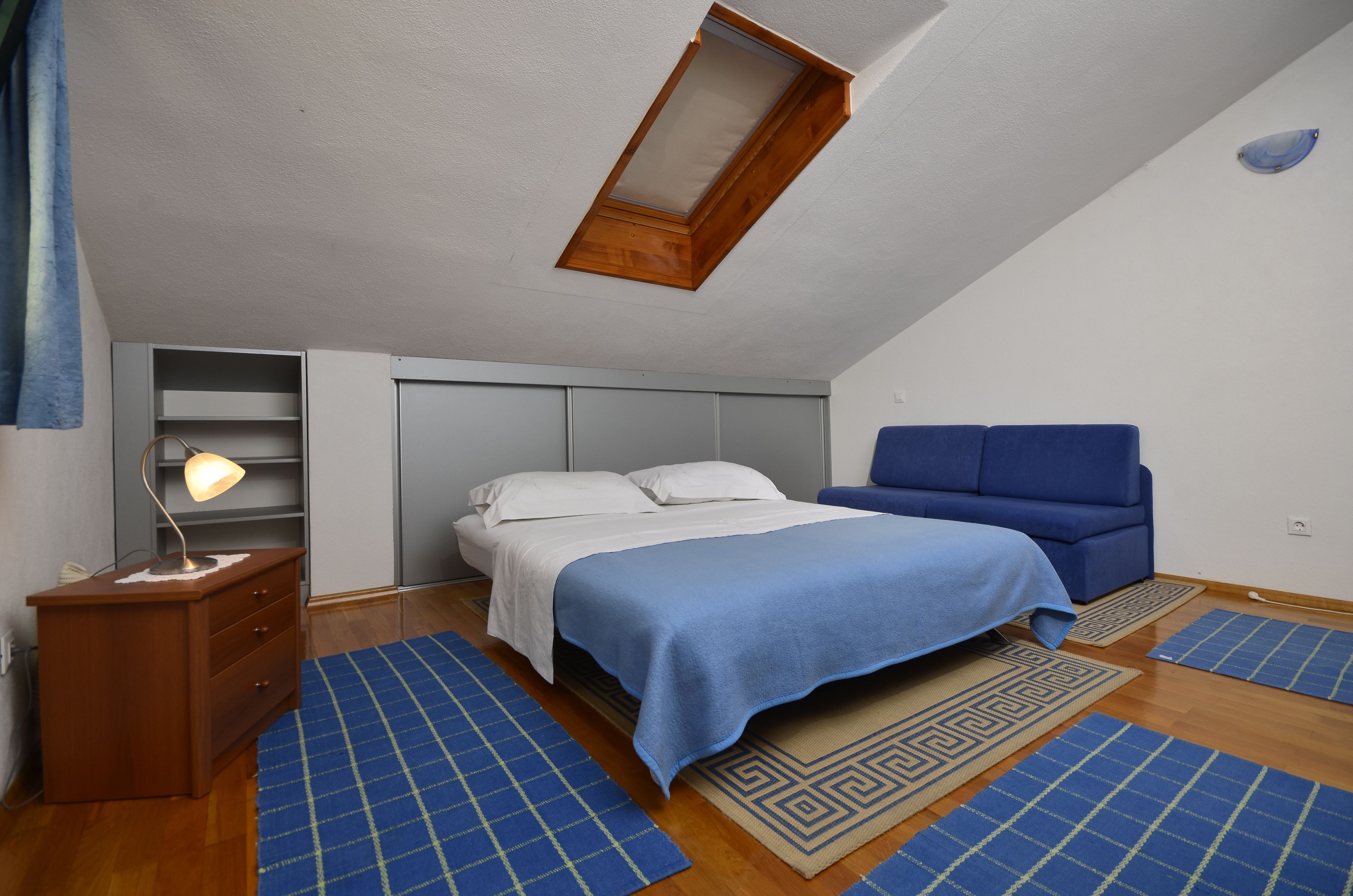 A bedroom with a bed, a blue sofa, a desk with a lamp, and a skylight.