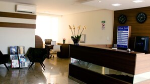 A hotel reception area with a wooden reception desk, two black chairs, a vase with flowers, a clock, and a sign listing room rates. - Hotel Belugi (Goiania)