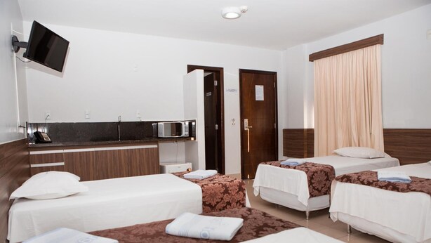 A hotel room with two single beds, a small kitchenette, a flat-screen TV, and a large window with curtains. - Hotel Belugi (Goiania)