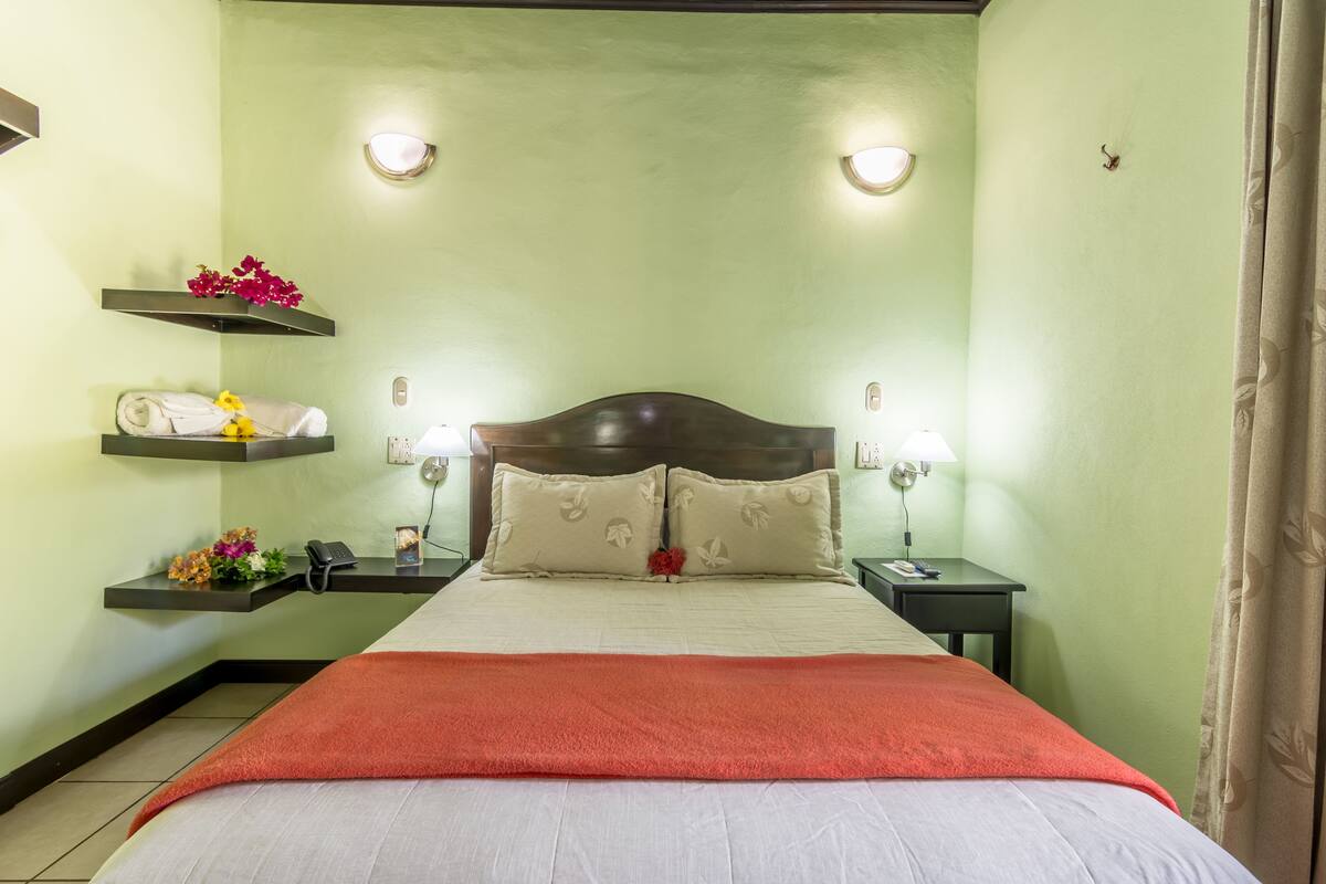 A neatly arranged hotel room with a bed, two bedside tables, a nightstand, and wall-mounted shelves with towels and flowers.