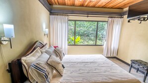 Ein Schlafzimmer mit einem großen Bett, einem Holzkopfteil, einem Nachttisch, einer Bank, einem an der Wand montierten Fernseher und einem Fenster mit Vorhängen.