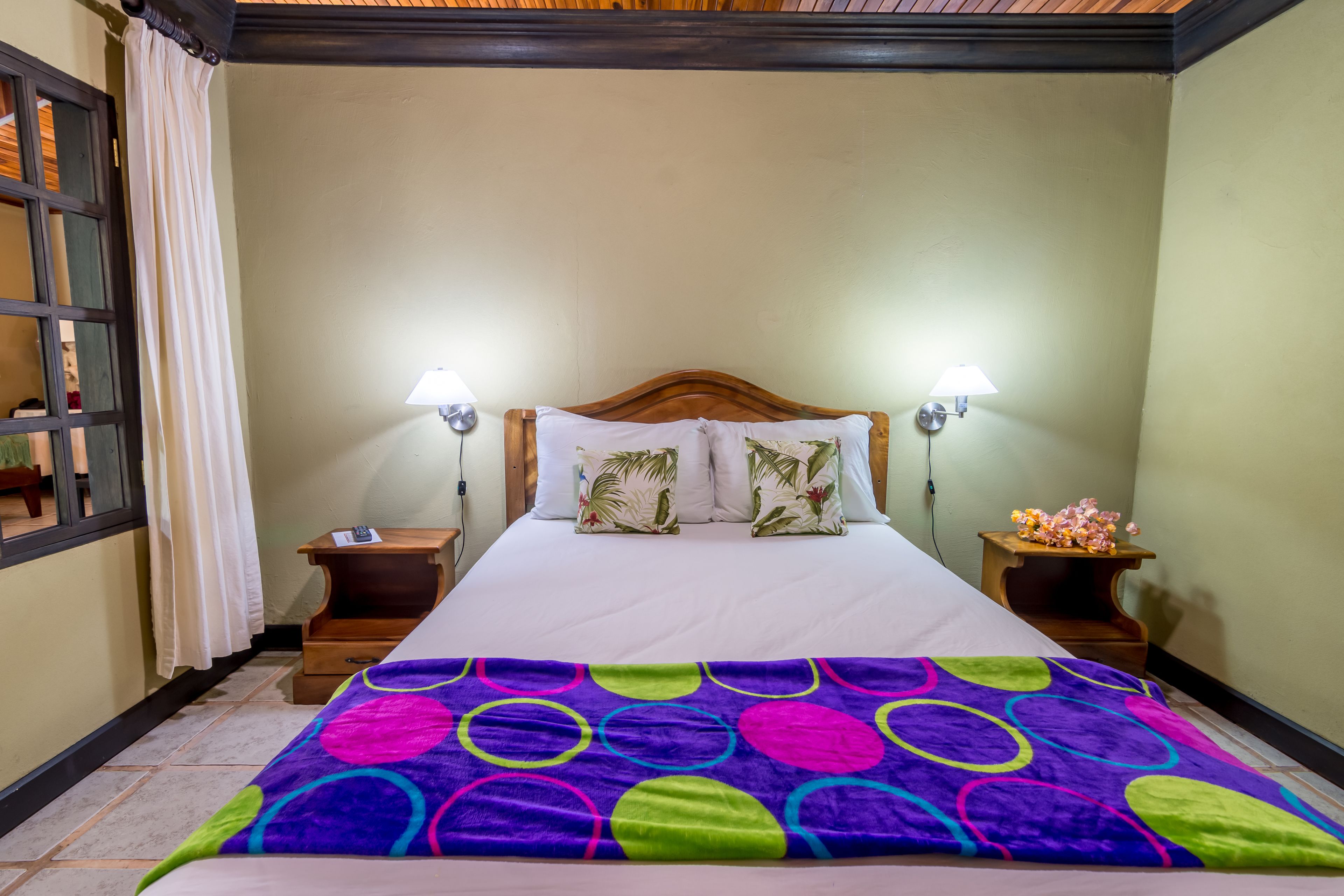 A neatly made bed with a colorful blanket, two bedside lamps, and a small table with a flower arrangement.