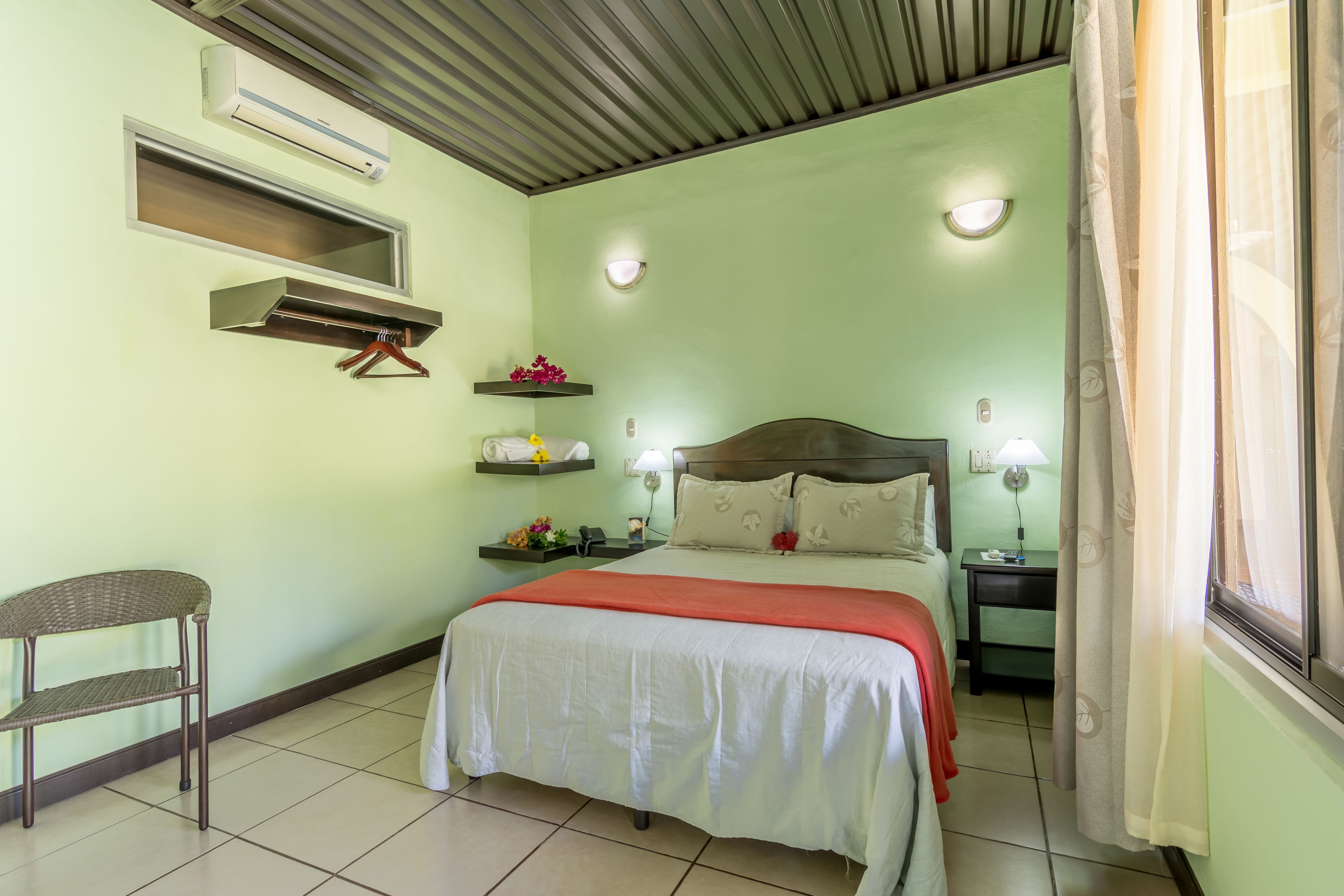 Quarto bem organizado com cama, mesas de cabeceira, uma cadeira e uma estante com flores.