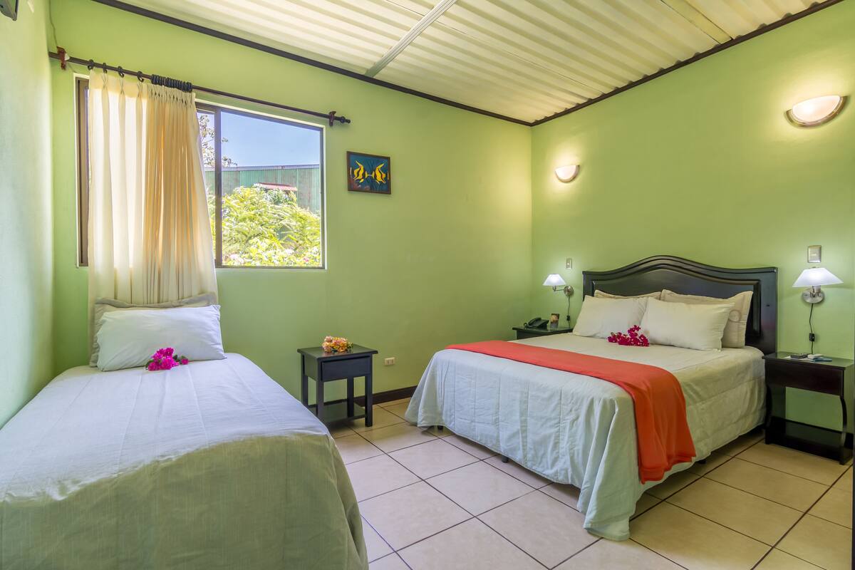 A room with two beds, a window with curtains, a small table with a lamp, and a painting on the wall.