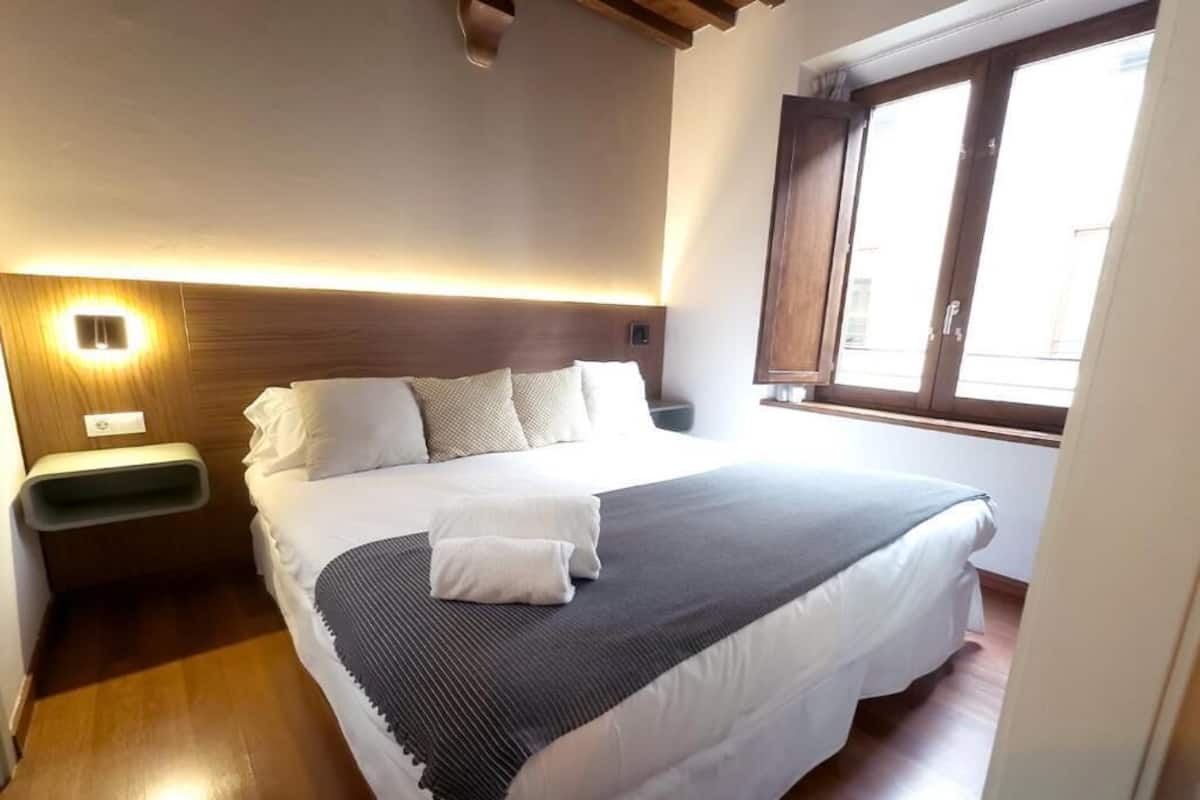 A neatly made bed with a white comforter and a gray blanket, a bedside table with a lamp, and a window with wooden shutters.