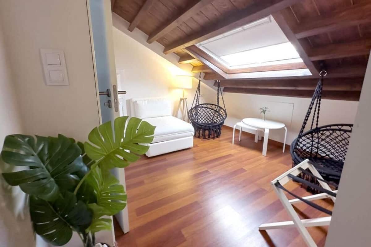 A cozy loft bedroom with a skylight, wooden beams, a bed, a hanging chair, and a small table.
