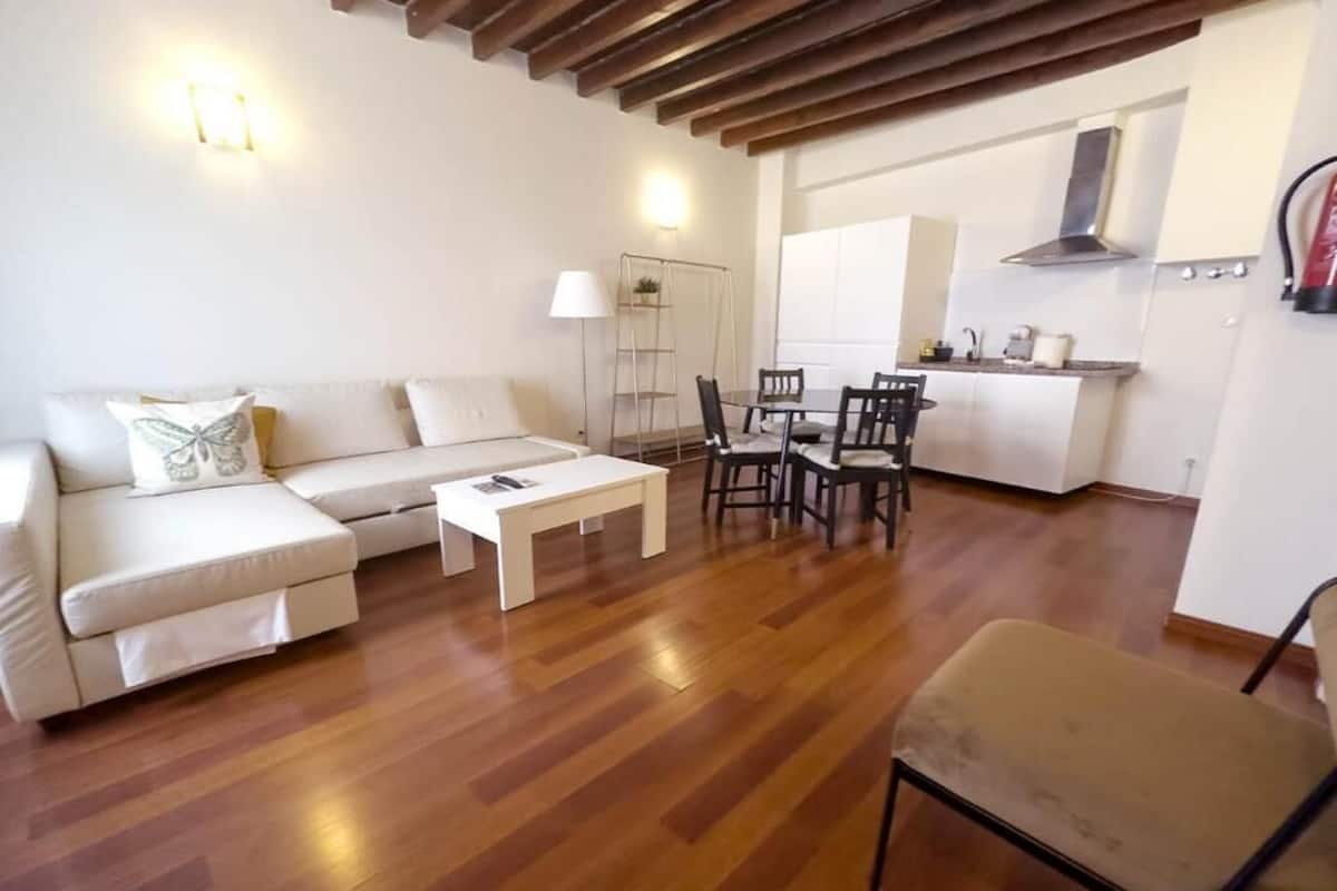 A living room with a sofa, coffee table, and chairs. There is a kitchen area with cabinets and a stove in the background.