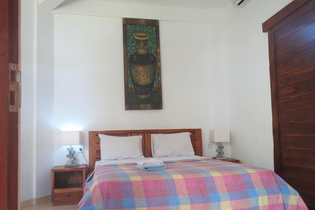Une chambre à coucher avec un lit en bois, une couvre-lit à carreaux colorés, une table de chevet avec une lampe et un tableau accroché au mur.