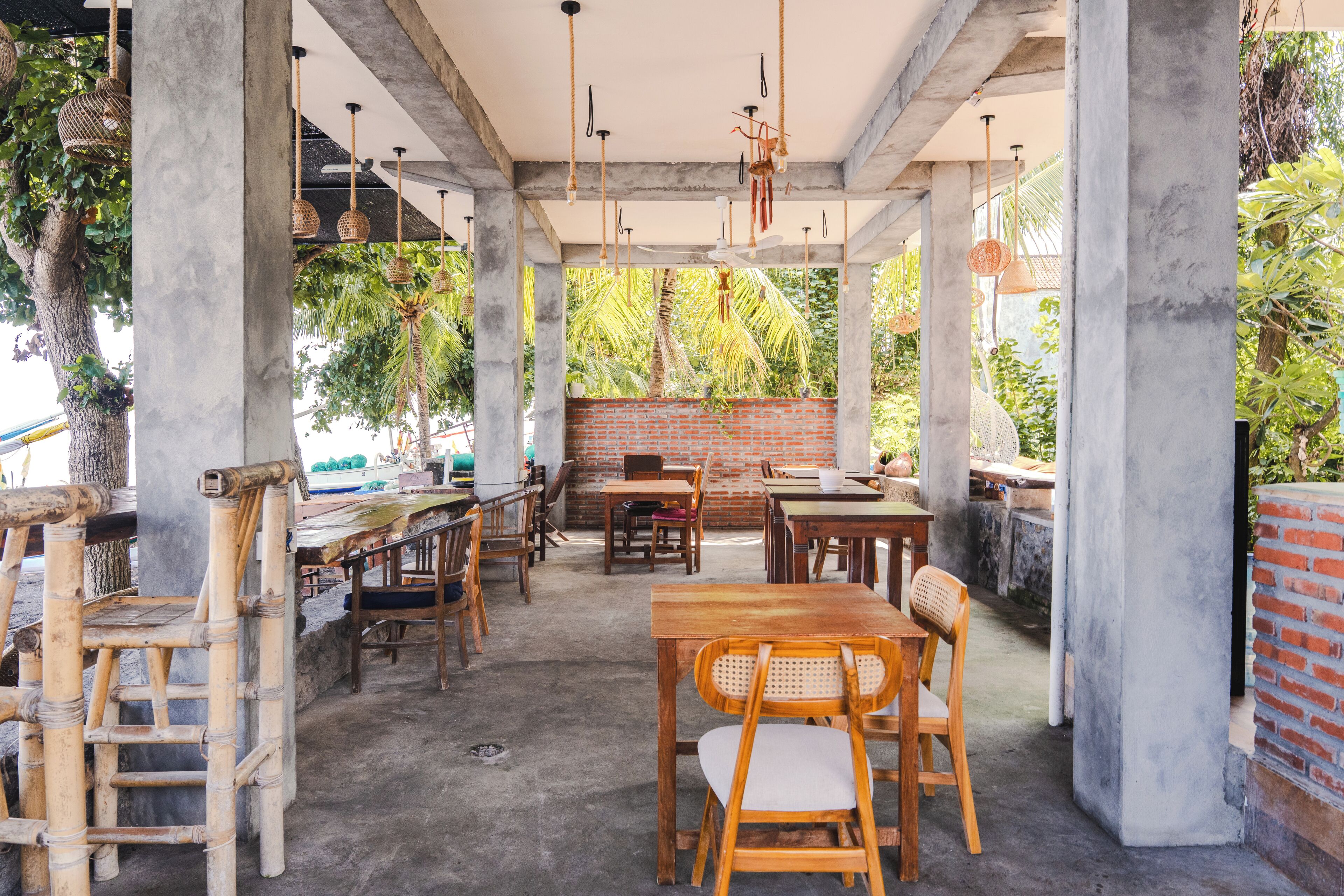 Un café al aire libre con pilares de hormigón, mobiliario de madera y luces colgantes.