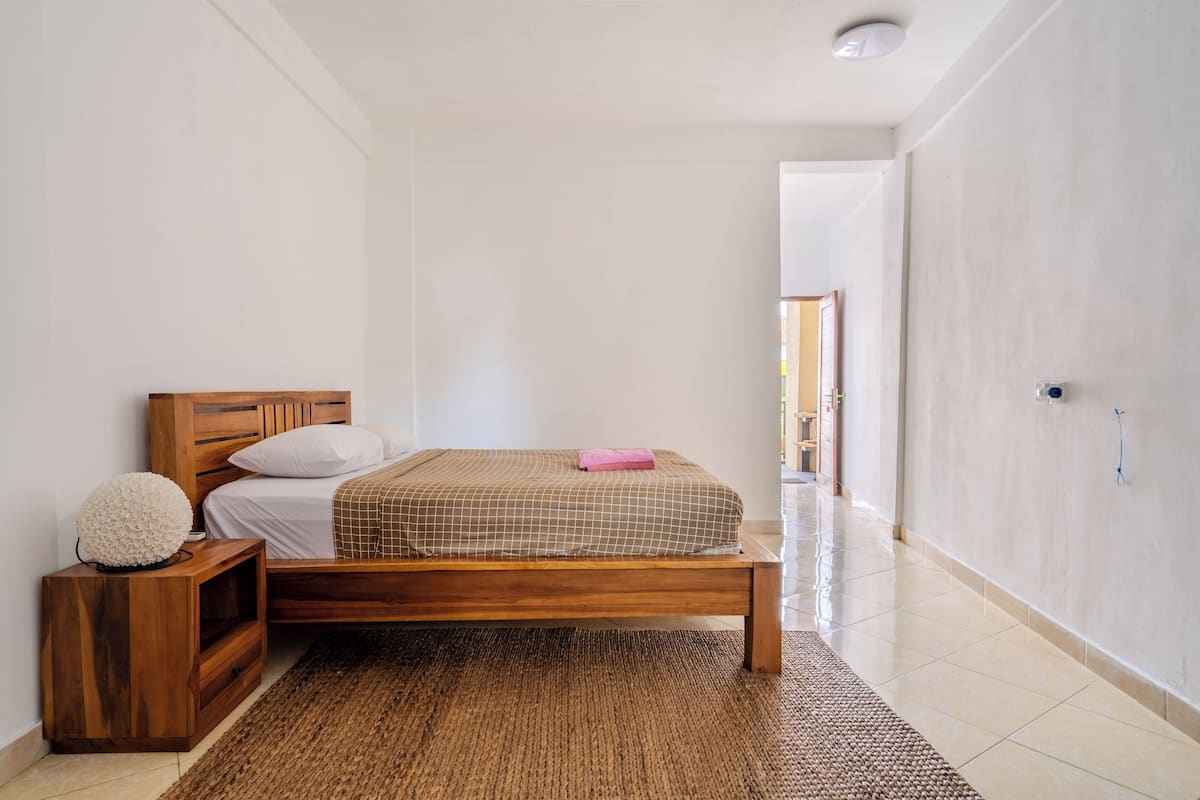 Une chambre avec un lit en bois, une table de chevet, un tapis tissé et un mur blanc.