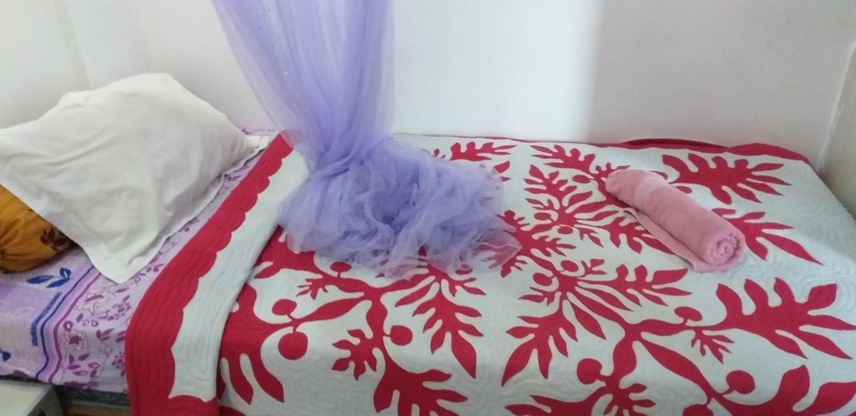 A bed with a red and white floral bedspread, a white pillow, and a rolled-up pink towel.